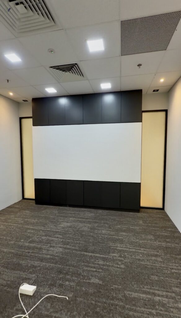 Empty room with grey carpet, white walls, ceiling lights, and a black and white panel on one wall; a power socket and lead are visible on the floor in the foreground.