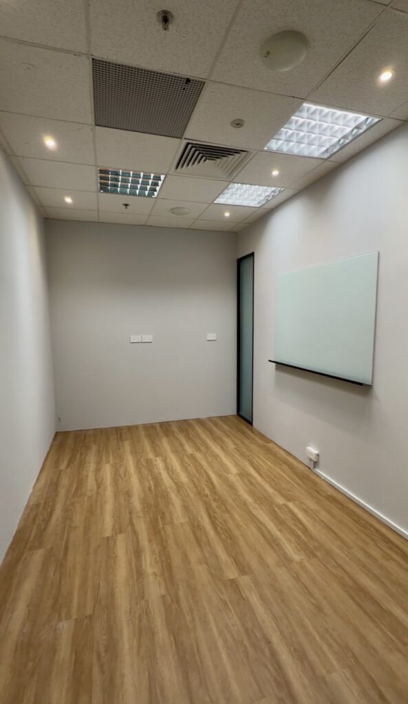 Empty office room with wood-look flooring, white walls, ceiling lights, a glass whiteboard on the right wall, and power sockets near the floor.