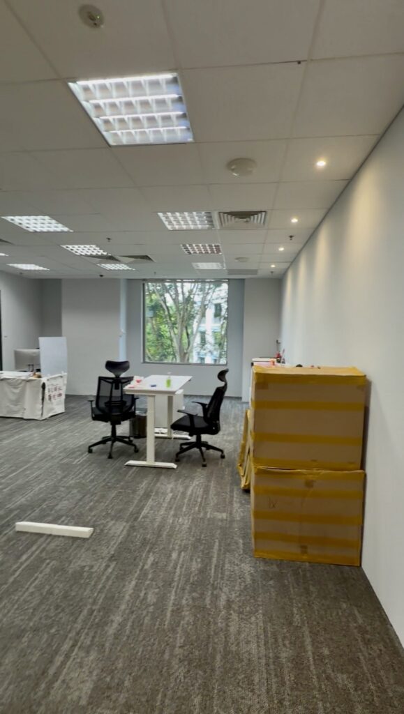 Office with two chairs at a desk, large boxes stacked against the wall, and a window showing trees outside.