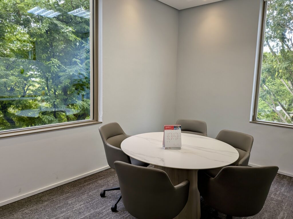 Small meeting room with a round table, four grey chairs, and two large windows showing green trees outside. A leaflet holder sits on the table.