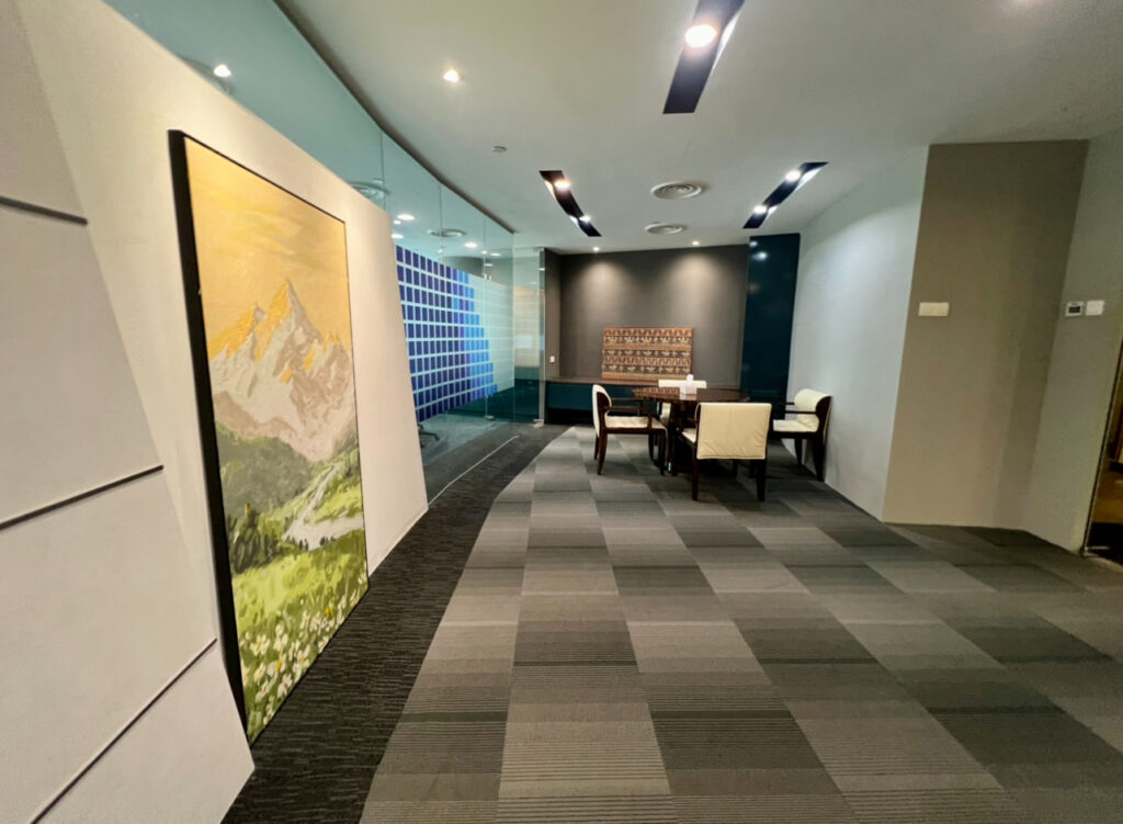 Modern office corridor with a mountain painting on the left, glass walls, and a small meeting area with four chairs and a table on the right.