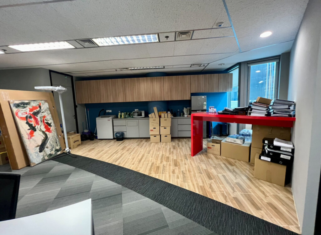 Office space with packed boxes, a large painting resting on the floor, a kitchenette area, and a red table with documents and office supplies stacked on top and underneath.