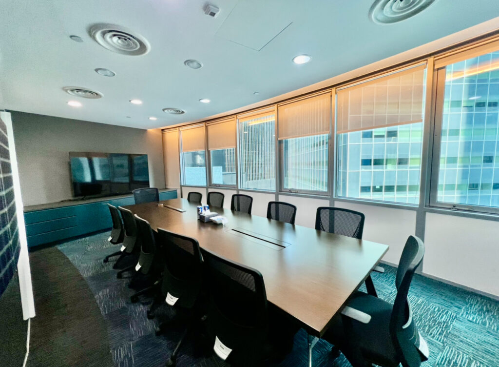 Modern conference room with a long table, ten black chairs, large windows, wall-mounted TV, and city buildings visible outside.