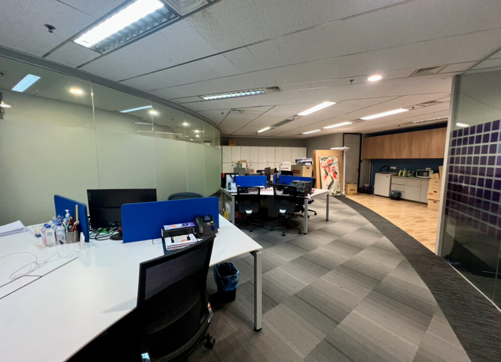 Modern office space with empty desks, computers, office chairs, and a glass wall; cupboards and artwork are visible in the background.