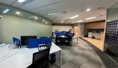 Modern office space with empty desks, computers, office chairs, and a glass wall; cupboards and artwork are visible in the background.