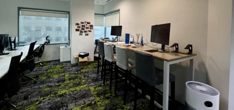 Modern office with desks, computers, chairs, and large windows. Wall with small photos, box on the floor, and an air purifier near two high-backed chairs on a green-patterned carpet.