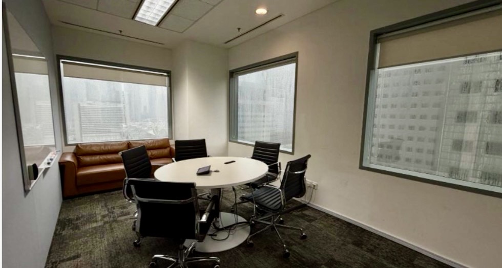 A small office meeting room with a round table, five chairs, a brown sofa, whiteboard, and large windows overlooking city buildings on a cloudy day.