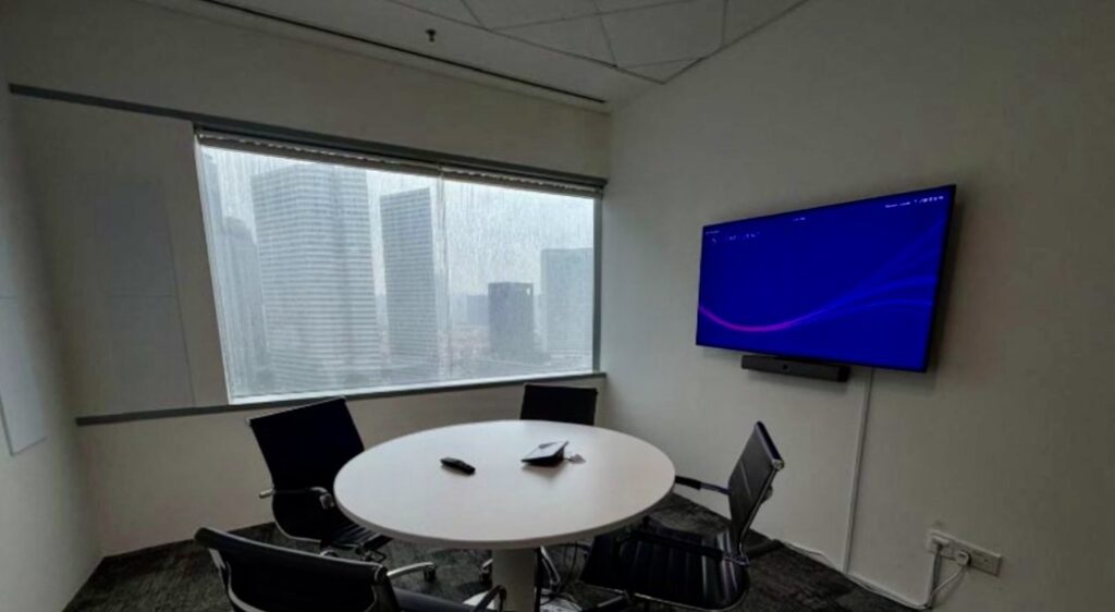 Small meeting room with a round table, four chairs, TV screen on the wall, window overlooking tall buildings, and remote controls on the table.