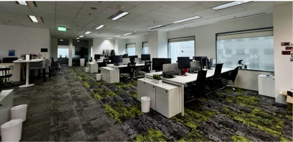 Modern open-plan office with multiple desks, computer monitors, chairs, and large windows. The room is empty, and the floor has a grey and green patterned carpet.
