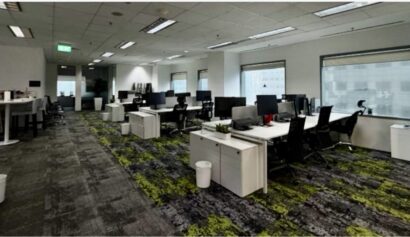 Modern open-plan office with multiple desks, computer monitors, chairs, and large windows. The room is empty, and the floor has a grey and green patterned carpet.