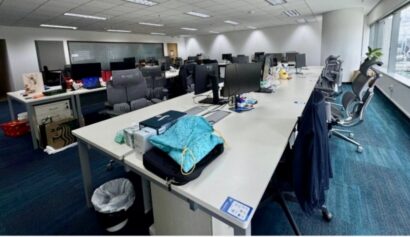 Open-plan office with empty desks, computer monitors, office chairs, scattered personal items, and large windows letting in natural light. The space appears organised and modern.