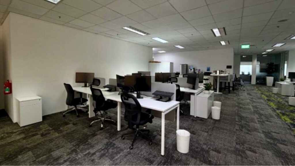 Modern open-plan office with multiple desks, computers, office chairs, and rubbish bins. The space is well-lit with overhead lights and has a grey carpeted floor.