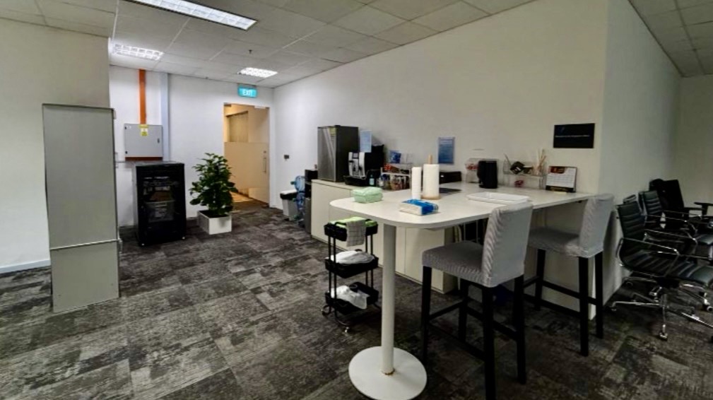 Office kitchenette area with a high table, two chairs, coffee machine, fridge, supplies on the worktop, and a row of chairs along the wall. A potted plant is near the corridor entrance.