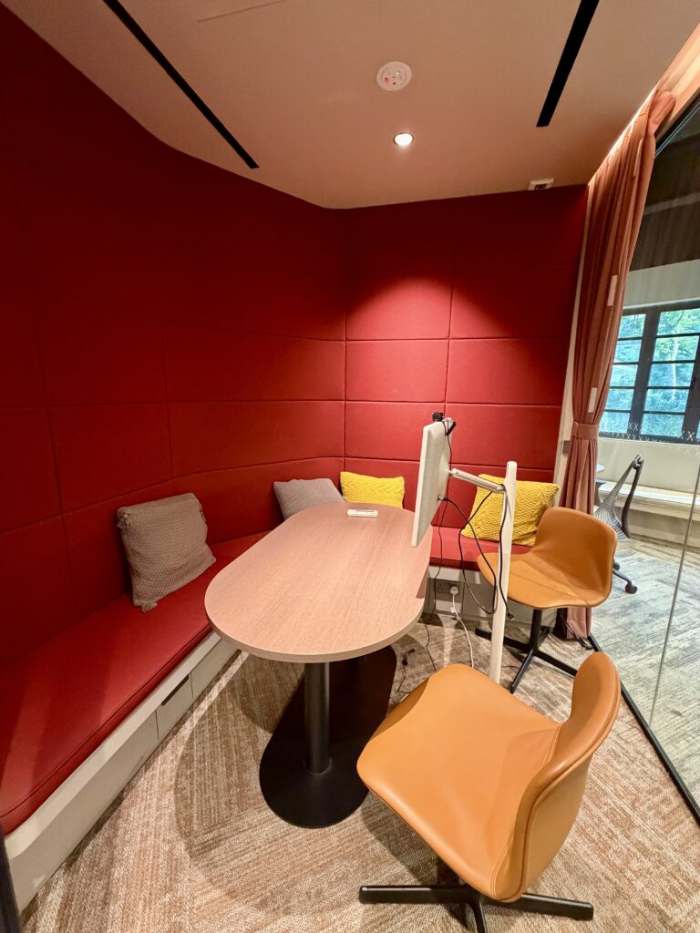 Small meeting room with red padded walls, a wooden table, orange and yellow chairs, a monitor on the table, and cushions on a built-in bench.