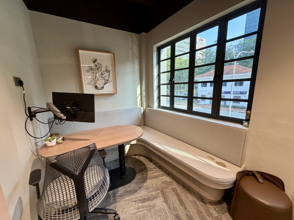 Small modern office nook with a desk, chair, mounted monitor, curved bench, window with city view, and framed artwork on the wall.