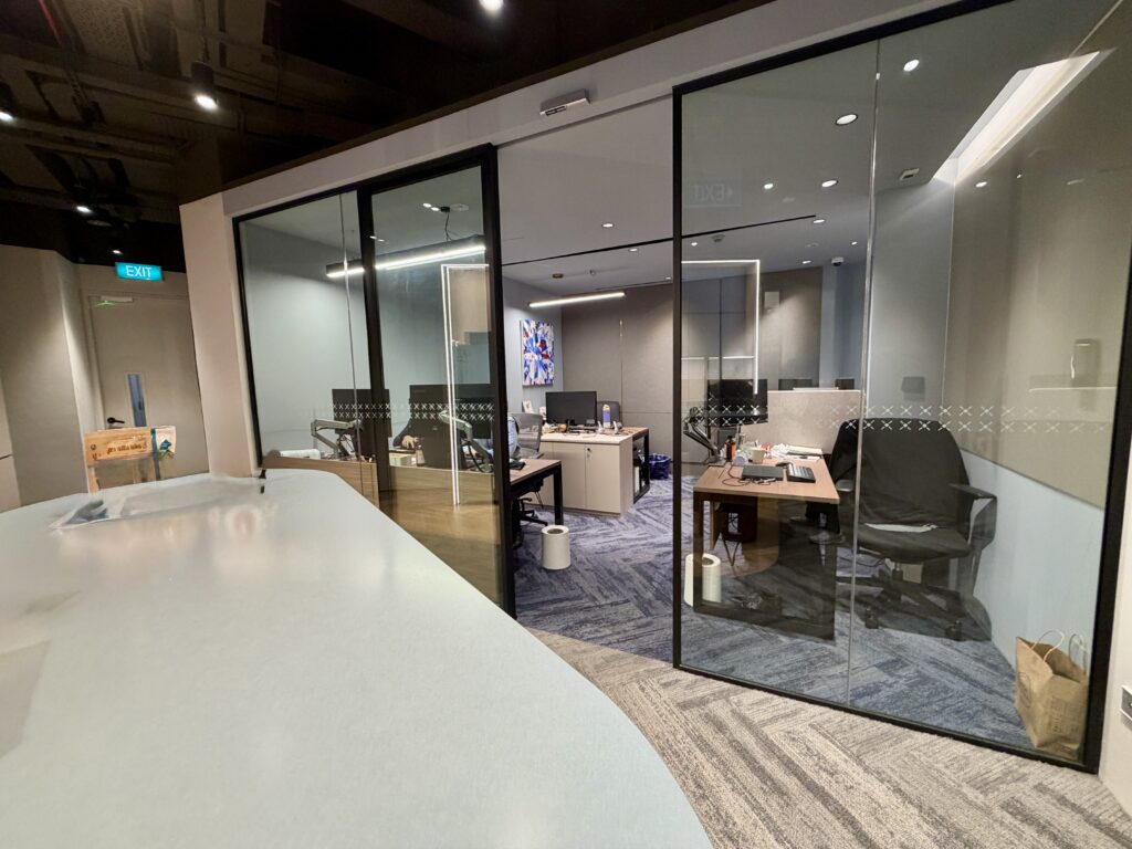 Modern office with glass-walled rooms, desks with computers, chairs, office supplies, and a wastepaper bin on a carpeted floor. A television screen is visible on the far wall.