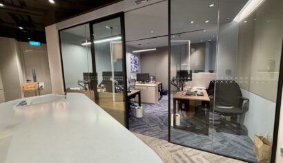 Modern office with glass-walled rooms, desks with computers, chairs, office supplies, and a wastepaper bin on a carpeted floor. A television screen is visible on the far wall.