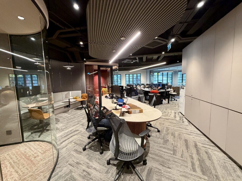 Modern open-plan office with several desks, chairs, computers, and office supplies. Glass-walled meeting room and large windows are visible in the background. Room is well-lit.