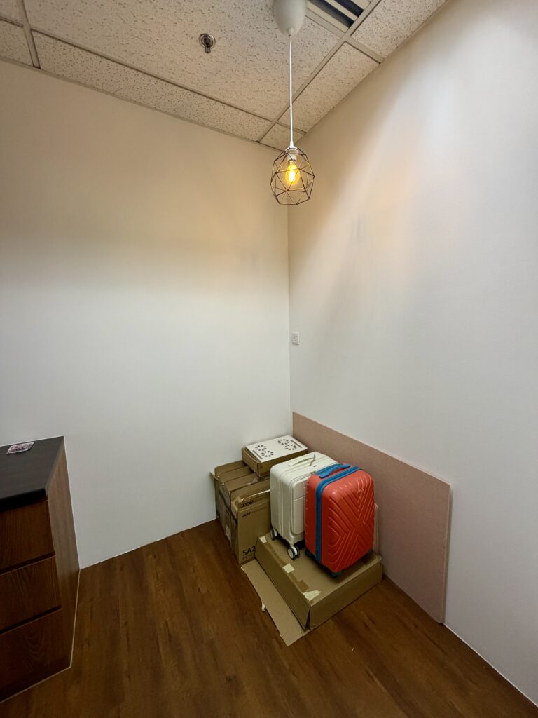 A small room with wooden flooring, white walls, a hanging light, some cardboard boxes, two suitcases, and an air cooler stacked in the corner.
