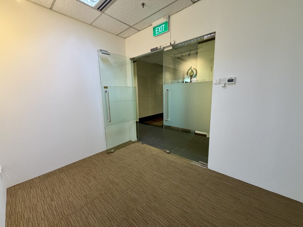 Open glass doors lead from a carpeted corridor into an office area with a visible exit sign above the doorway.
