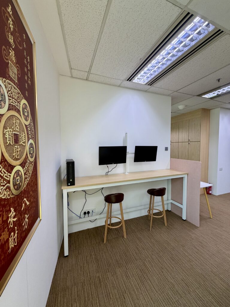 Two stools and computer monitors at a long desk in a modern office space, with a large red decorative wall hanging on the left and brown carpeted floor.