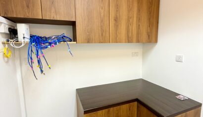 Office corner with L-shaped wooden desk, upper cupboards, and exposed blue network cables hanging from an open compartment on the left wall.