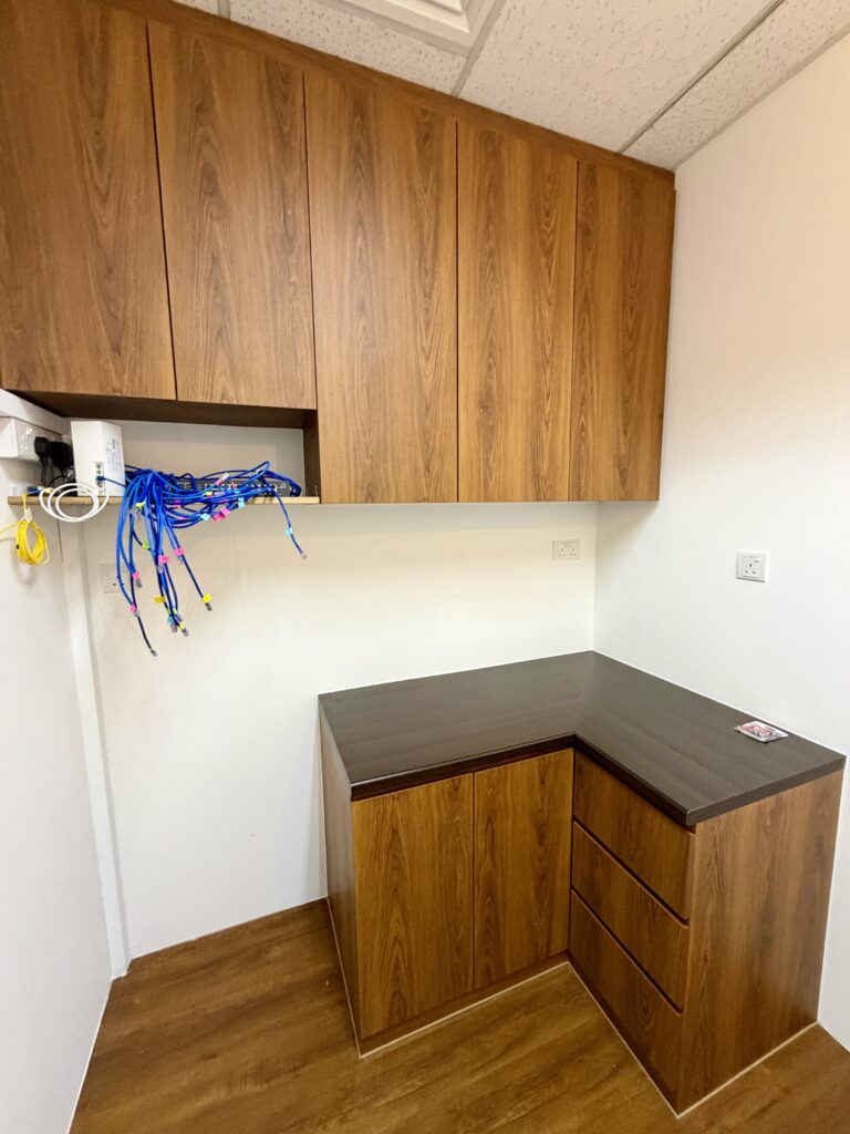 Office corner with L-shaped wooden desk, upper cupboards, and exposed blue network cables hanging from an open compartment on the left wall.