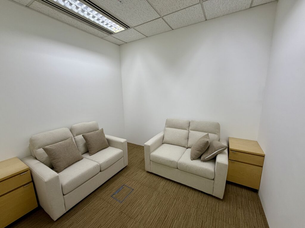 Small office room with two beige sofas, four brown cushions, two wooden side tables, white walls, and carpeted floor under fluorescent lighting.