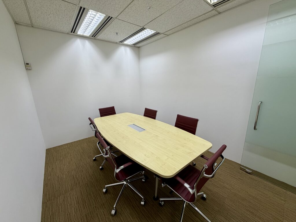 Small meeting room with a rectangular table, six red chairs, beige carpet, white walls, and a glass door.