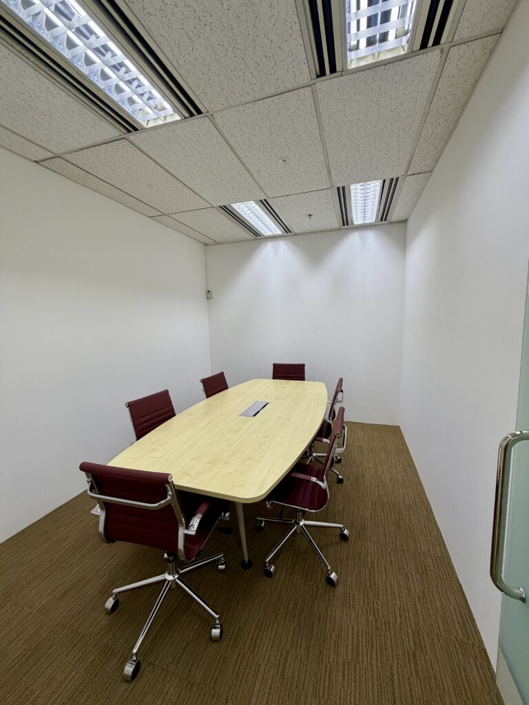 A small meeting room with a long wooden table, six red office chairs, white walls, and fluorescent ceiling lights.