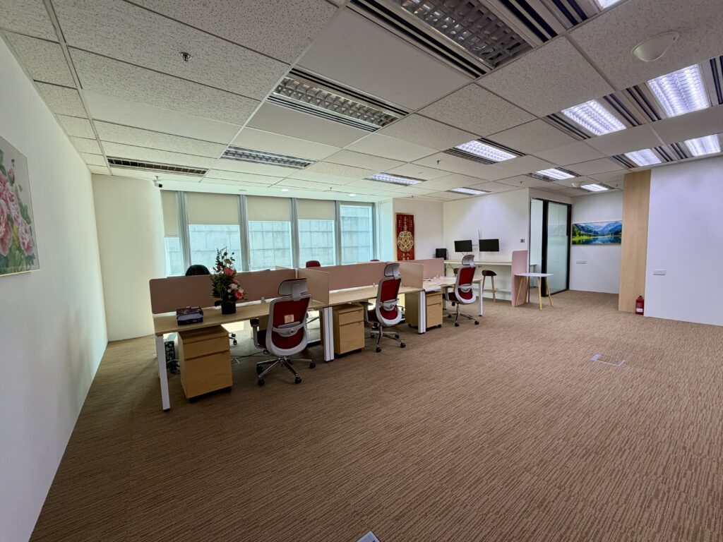 Modern office space with several workstations, chairs, computers, and decor including plants and wall art. The room is well-lit with fluorescent lights and has beige carpeting.