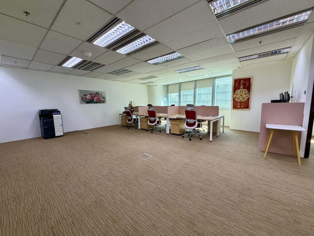 Spacious office with four desks and chairs, a printer, wall art, and large windows letting in natural light. A small table stands near the right wall.