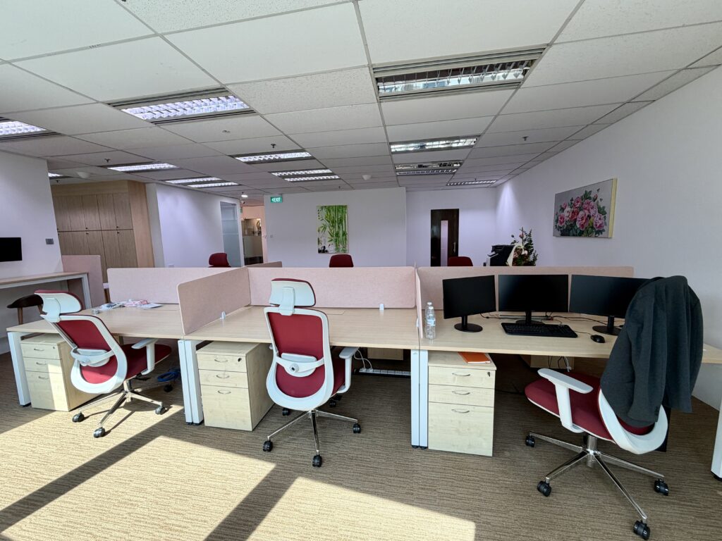 Modern open-plan office space with four desks, red and white chairs, computer monitors, a water bottle, and flower artwork on the wall. Brightly lit by overhead lighting and sunlight.