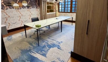 Modern office with a large table, blue patterned rug, wooden floors, geometric wall design, large windows, and pendant lighting.