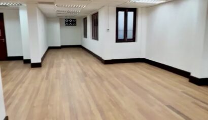 Empty office space with white walls, wooden flooring, fluorescent ceiling lights, windows on the right, and a door at the far end.