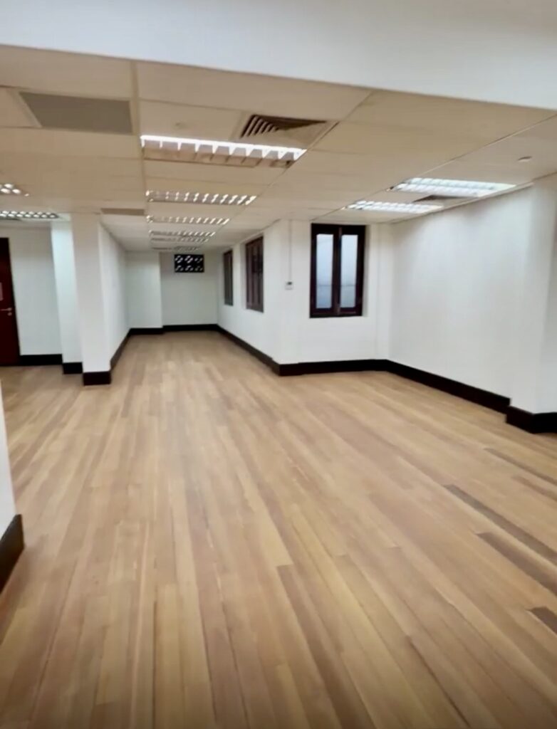 Empty office space with white walls, wooden flooring, fluorescent ceiling lights, windows on the right, and a door at the far end.