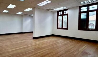 Empty room with wooden floor, white walls, ceiling lights, and two windows letting in natural light.