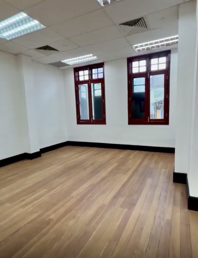 Empty room with wooden floor, white walls, two red-trimmed windows, and ceiling lights.