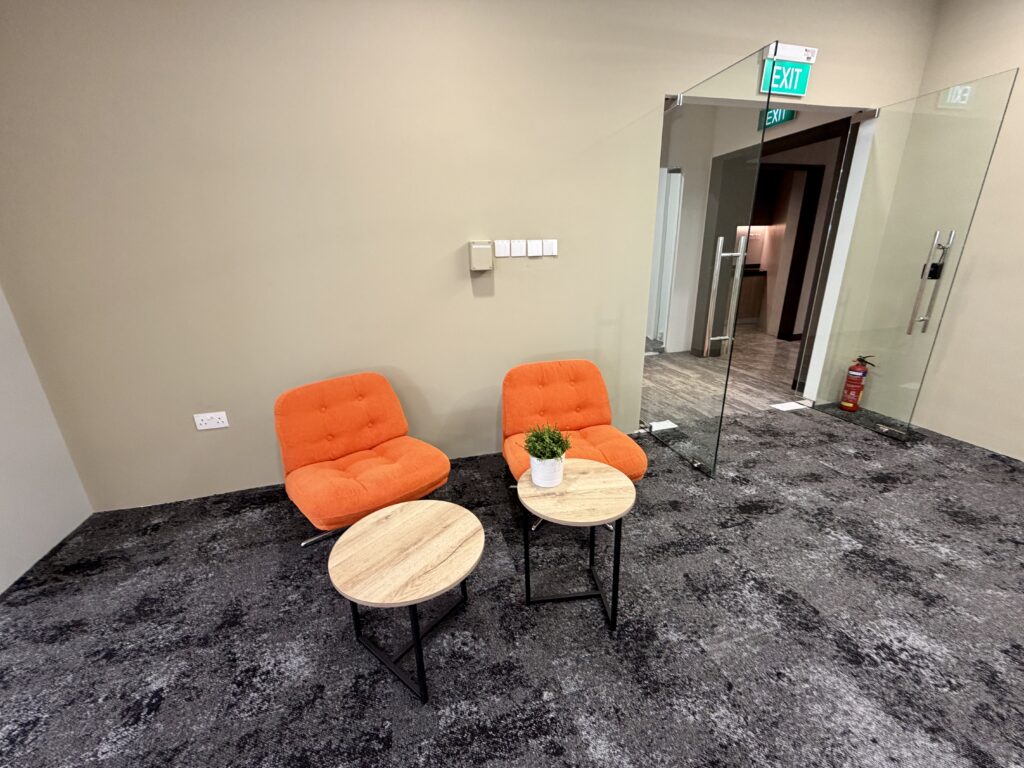 Two orange chairs and two round wooden tables sit on a grey carpet near a beige wall. A small potted plant is on one table. Glass doors and an exit sign are visible in the background.