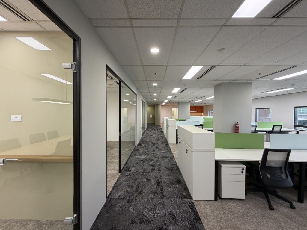 Modern office corridor with glass-walled meeting rooms on the left and open-plan workstations with chairs and dividers on the right. Carpeted floor and ceiling lights are visible.