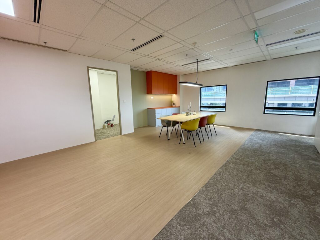Modern office space with a table, six colourful chairs, overhead lighting, and a kitchenette in the background; windows show exterior buildings.
