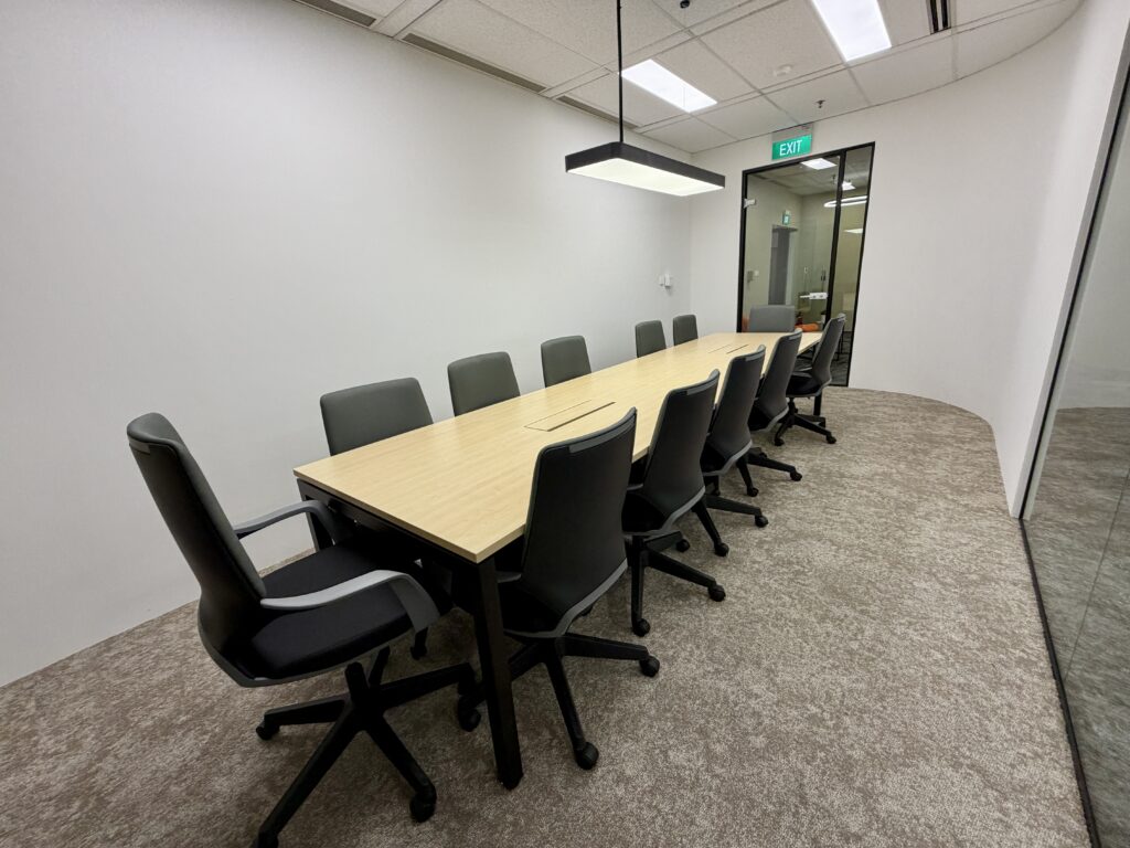 A conference room with a long wooden table, ten office chairs, beige carpet, white walls, a ceiling light, and a glass door with an exit sign visible.