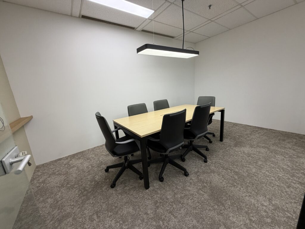 A small meeting room with a rectangular table, six black office chairs, white walls, carpeted floor, and overhead lighting.