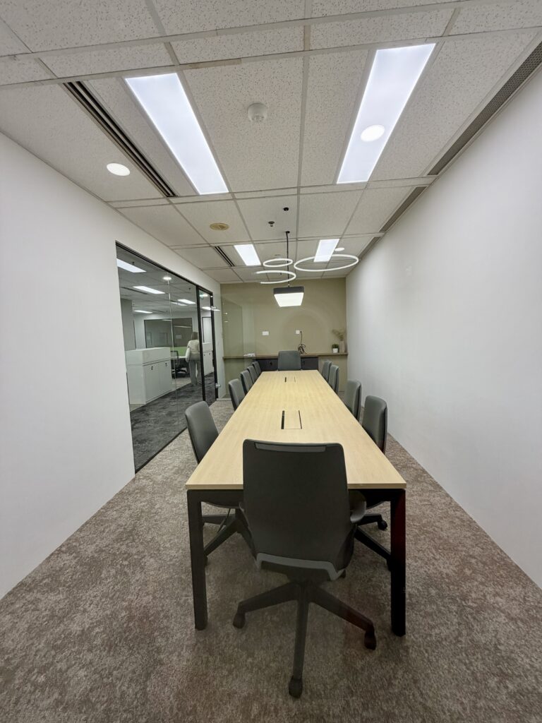 Long rectangular meeting room with a wooden table, eight chairs, white walls, carpeted floor, glass wall on one side, and circular ceiling lights.