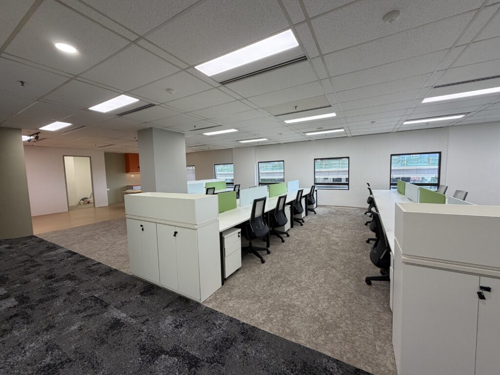 Modern open-plan office with rows of empty desks and chairs, carpeted floor, overhead lighting, and large windows letting in natural light.