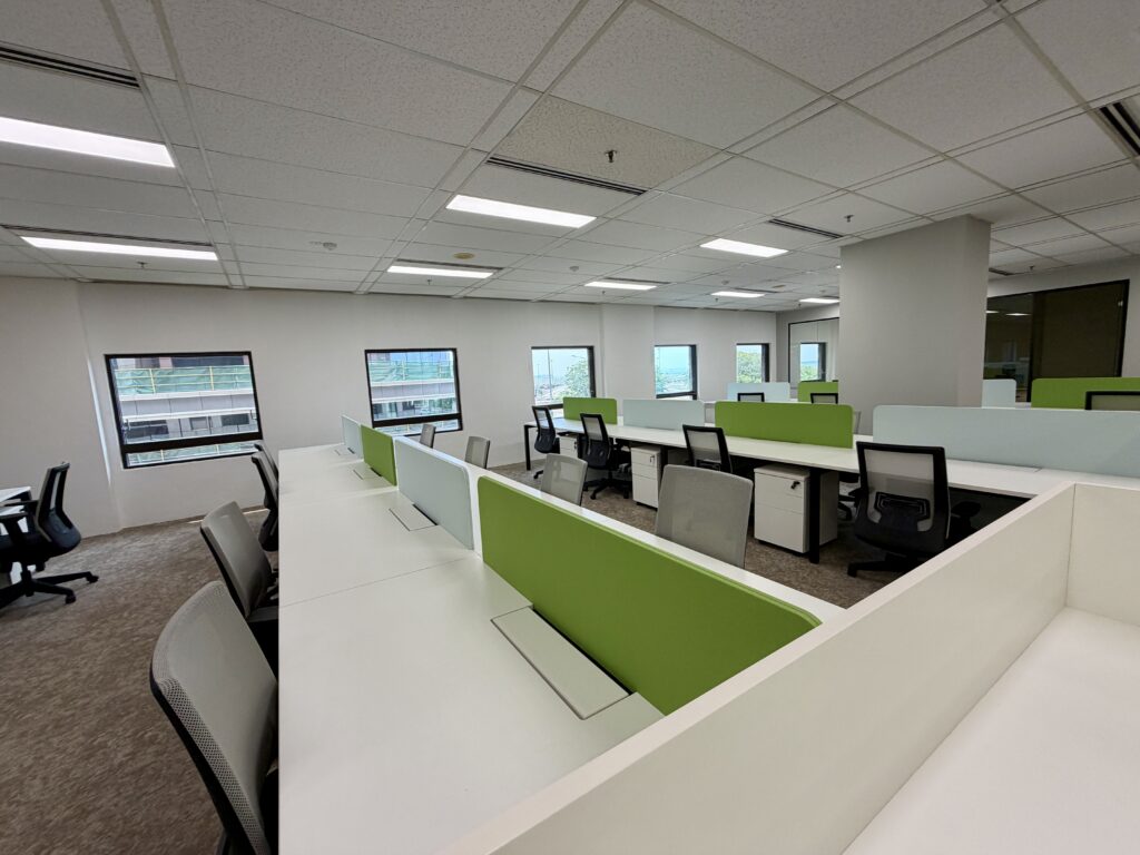 Modern office space with rows of empty desks and chairs, green partitions, large windows, and overhead fluorescent lighting.
