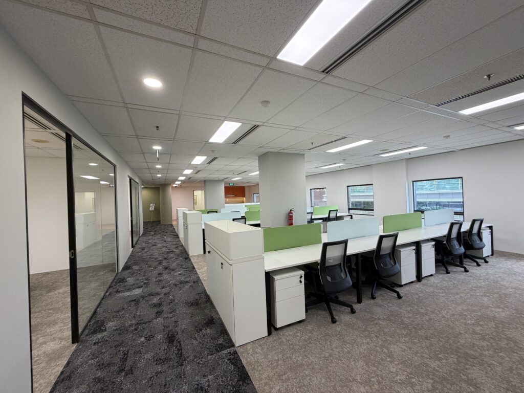 Modern open-plan office with rows of white desks and black chairs, green dividers, large windows, and grey carpeting under bright overhead lights. No people are present.