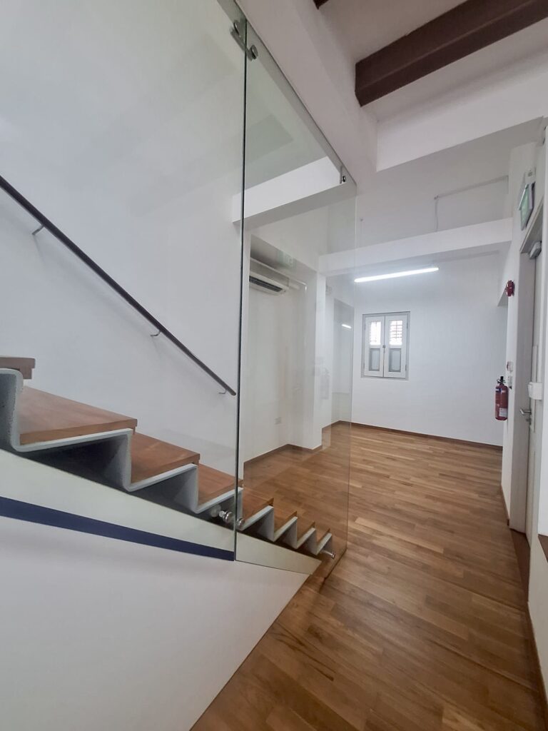 Interior view of a modern hallway with wooden floors, a glass stair balustrade, stairs on the left, and white walls.