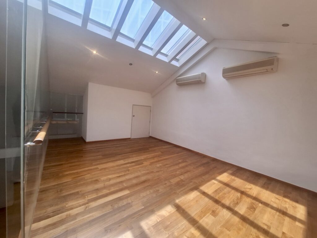 A spacious room with wooden floor, white walls, two wall-mounted air conditioners, a skylight ceiling, and a door at the far end. Sunlight streams in through the skylights.