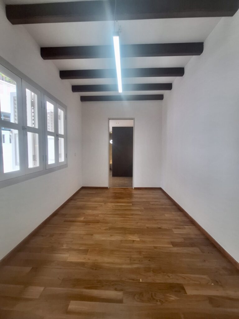 Empty room with wooden floor, white walls, exposed dark ceiling beams, a row of windows on the left, and a hanging fluorescent light.